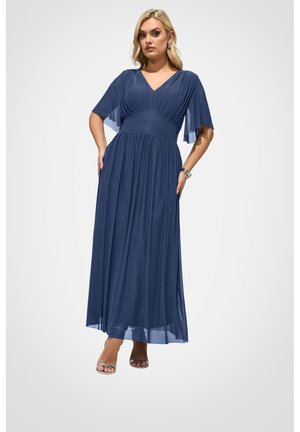 Frau posiert in einem bodenlangen marineblauen Kleid mit plissiertem Taillenbereich und flatternden Ärmeln, trägt silberne High-Heel-Sandalen und ein Armband.