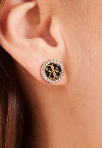 Gouden studoorbel met een zwarte centrale schijf waarop verweven initialen zijn afgebeeld, omringd door heldere kristallen in een ronde zetting.