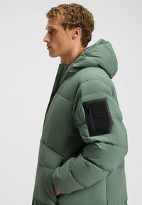 Gepolsterte Parka in gedämpftem Grün, mit Kapuze, Reißverschlusstasche am linken Arm und genoppter Textur zur Isolierung.