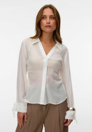 Femme portant un chemisier blanc transparent boutonné avec des poignets noués et un pantalon beige taille haute sur un fond uni.