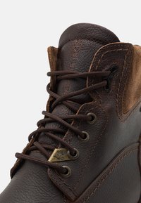 Bota de cuero marrón con acentos de gamuza, cordones redondos, ojales de metal y logo grabado. Presenta superficies texturizadas y costuras reforzadas.