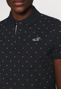 Svart polo skjorta med vitt prickmönster, kortärmad och med krage. Har en knappställd halsringning och en liten broderad logga.