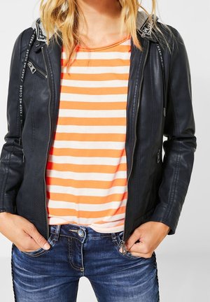 Frau trägt eine schwarze Lederjacke über einem orange-weiß gestreiften Shirt, kombiniert mit blauen Jeans, Hände in den Taschen.