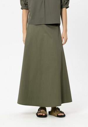 Unterer Teil einer Person, die einen olivgrünen A-Linien-Rock, ein passendes Oberteil und olivgrüne Sandalen mit Schnallen und Nieten trägt.