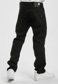 Person med svarte jeans og hvite Nike Air-sko, med ryggen til, står mot en enkel lys bakgrunn.
