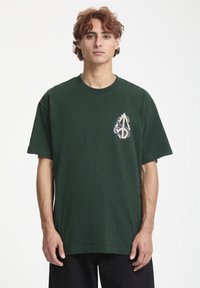 Grøn bomulds T-shirt med korte ærmer, rund halsudskæring, prydet med et lilla og cremefarvet grafisk design på venstre brystområde.