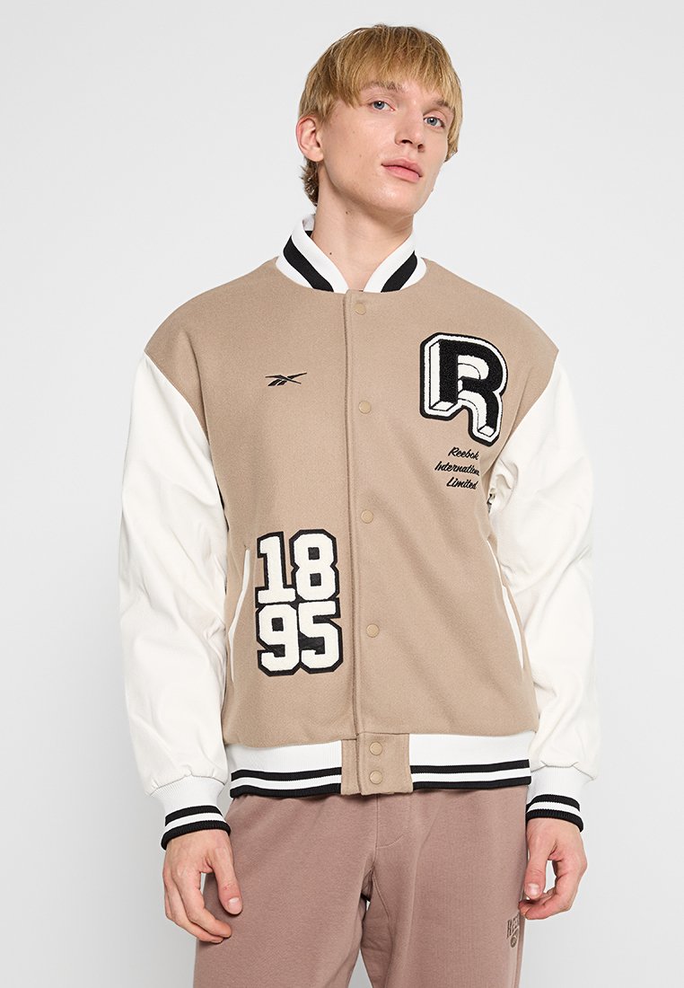 Veste universitaire Reebok, beige et blanche, avec des accents noirs, des lettres et des chiffres brodés, des boutons-pression et des poignets côtelés.