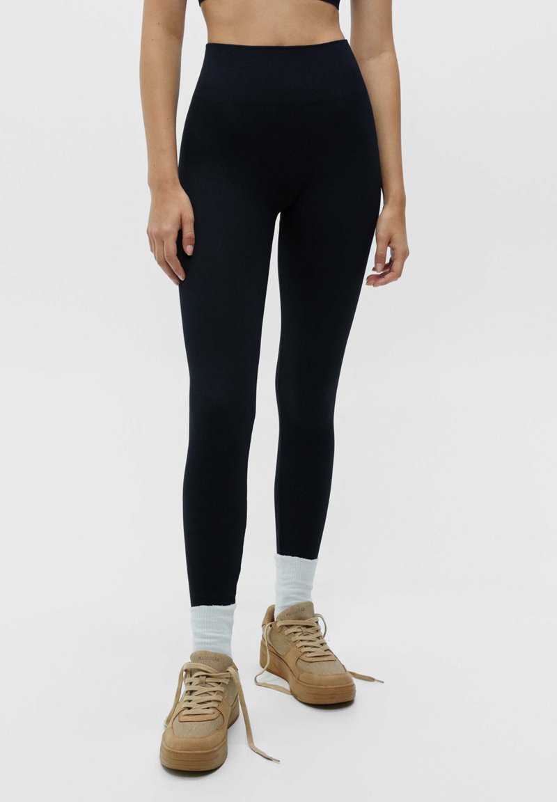 Leggings noirs taille haute en matière extensible, avec une texture lisse et une coupe ajustée, accompagnés de chaussettes bleu clair et de baskets beige.