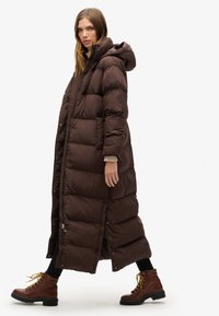 Brun pufferjakke med et langt, oversized design, der har hætte, trykknaplukninger og sidelommer, parret med brune snørestøvler.