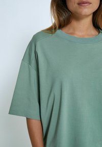 Světle zelené oversized tričko s krátkými rukávy, kulatým výstřihem a měkkou bavlněnou tkaninou. Má jemné prošití podél rukávů a lemů.