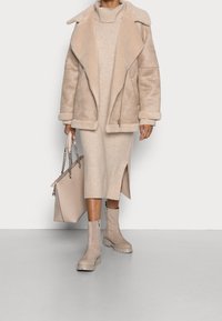 Veste en fausse peau de mouton beige avec grand col, robe beige en tricot avec fente latérale et bottines beiges à semelles épaisses. Porte un sac fourre-tout.