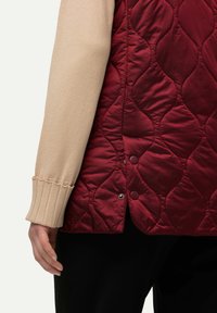 Quiltny bordowy kamizelka z gładką, błyszczącą fakturą i okrągłymi zatrzaskami, w połączeniu z beżowym swetrem z prążkowanego materiału. Czarne spodnie widoczne poniżej.