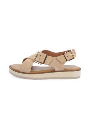 Sandale plate en daim beige avec des brides croisées, deux boucles ajustables dorées, une semelle intérieure rembourrée et une semelle plateforme blanche.