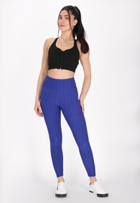Černý crop top se zipem vpředu a širokými ramínky, kombinovaný s texturovanými modrými legínami s hexagonálním vzorem, obutý s bílými teniskami.