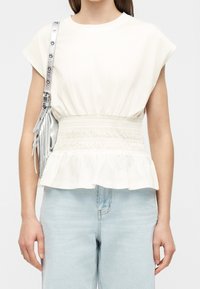Femme portant un chemisier blanc à manches courtes avec taille froncée et un jean bleu clair, portant un sac à épaule argenté avec des franges.