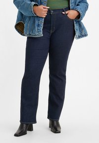 Jeans de ganga escuros de perna reta com bolsos cosidos, cintura alta e costuras subtis, combinados com uma blusa de listas verdes e uma jaqueta azul oversized.