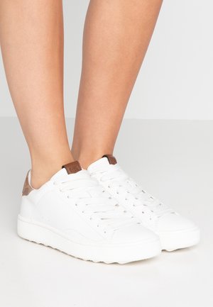 Zapatillas blancas bajas con detalles marrones usadas en los pies sobre un fondo claro y liso, mostrando vistas laterales y frontales.