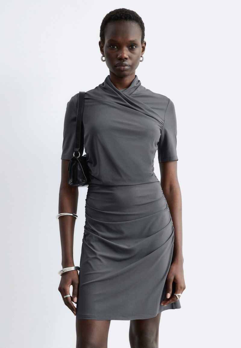 Femme portant une robe grise cintrée à manches courtes avec une jupe froncée, portant un sac à épaule noir et des bijoux en argent.