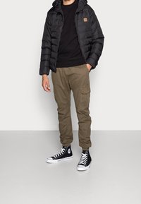 Doudoune noire à capuche, sweat-shirt noir en dessous, pantalon cargo olive, et baskets montantes noires avec des accents blancs.