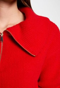 Rode gebreide trui met een geribbelde textuur, voorzien van een hoge kraag en een goudkleurige rits aan de voorkant.