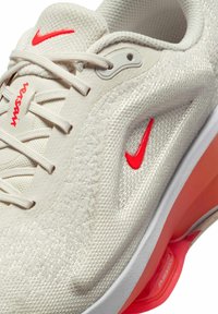 Bežs sporta apavu modelis ar teksturētu augšdaļu, kuram ir sarkans akcents uz Swoosh logo un redzams oranžs Air Zoom amortizators zoles daļā.