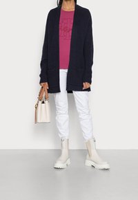 Cardigan a coste blu navy con tasche, t-shirt grafica rosa, pantaloni bianchi e stivaletti alla caviglia con suola spessa bianchi. Borsa beige con manici marroni.