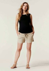 Top preto sem mangas combinado com calções bege claro com cintura ajustável. A modelo está de pé com as mãos nos bolsos, usando sandálias planas.