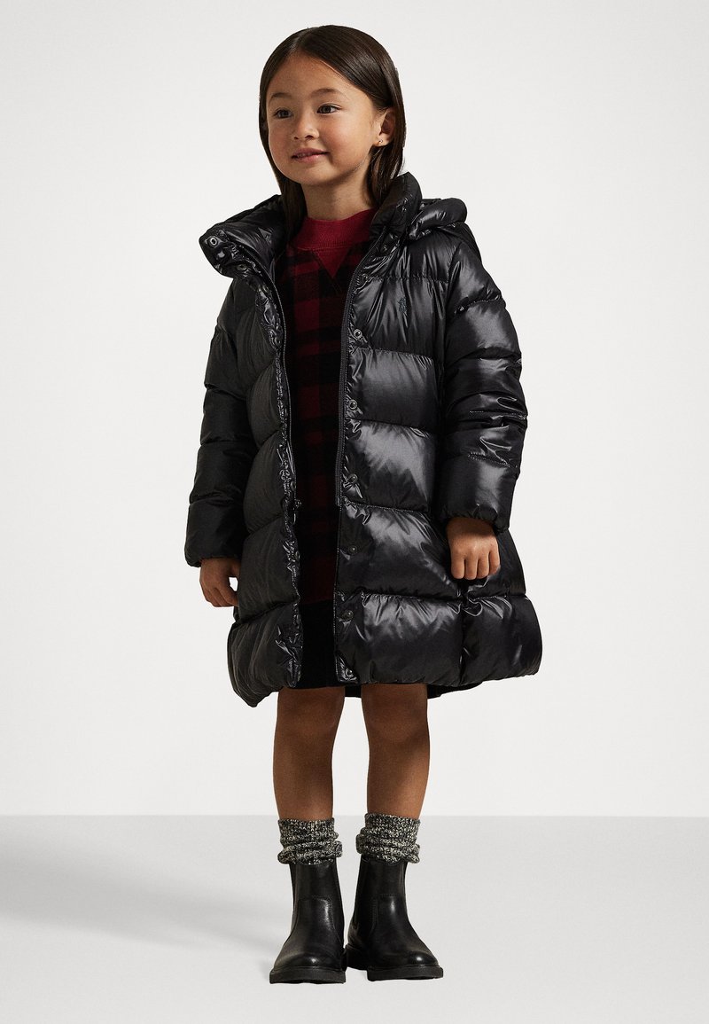 Casaco puffer preto e volumoso com capuz, apresentando uma textura brilhante e grandes secções acolchoadas. Usado sobre um vestido de xadrez vermelho e botas pretas.