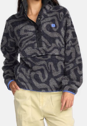 Mujer con un forro polar de color gris oscuro con estampado, botones de presión negros y pantalones beige, una mano en el bolsillo, torso visible.