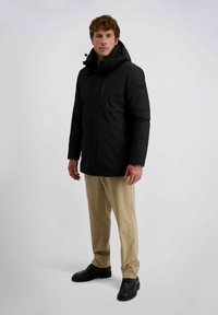 Schwarze isolierte Parka mit Kapuze, Frontreißverschluss und Seitentaschen; getragen mit hellbeigen Hosen und schwarzen Lederschuhen.