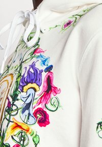 Sudadera blanca con gran bordado floral colorido que presenta flores moradas, rosas, azules y amarillas entrelazadas con enredaderas verdes.