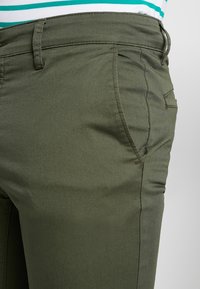 Spodnie w oliwkowej zieleni o gładkim, teksturowanym wykończeniu, z bocznymi kieszeniami i dopasowanym krojem, przeszywane nicią w tym samym kolorze.