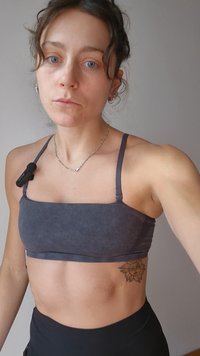 Gray athletic crop top with thin straps, smooth texture, featuring a sunflower tattoo on the lower abdomen and a necklace.