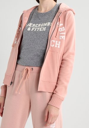 Kobieta ubrana w szarą koszulkę Abercrombie & Fitch pod różową bluzą na zamek i pasujące różowe spodnie dresowe.