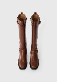 Une paire de bottes en cuir marron, hautes, avec des bouts carrés, des zips latéraux et des boucles dorées décoratives, photographiée sur un fond blanc.