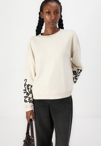 Krem sweatshirt z črnim abstraktnim vezenjem na rokavih, s sproščenim krojem in rebrastimi manšeti, kombiniran s temnimi kavbojkami.