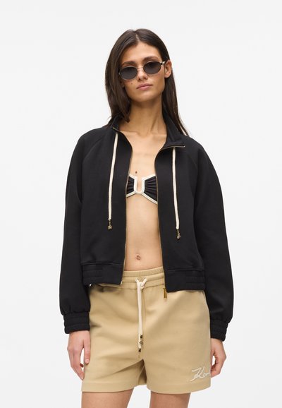 Veste noire zippée avec cordons et poignets côtelés, portée sur un haut de bikini noir et blanc, associée à un short beige à cordon de serrage.