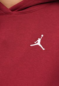 Hvit brodert basketballspillerlogo med utstrakt arm på burgunder hettegenserstoff nær halslinjen.