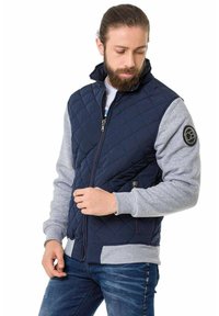 Marineblau gesteppte Jacke mit grauen Ärmeln und Kragen, mit durchgehendem Reißverschluss, seitlichen Taschen und einem runden Logo-Patch am linken Arm.
