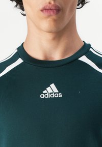 Mørkegrønn trenings T-skjorte med hvite accentstriper på skuldrene og en hvit brodert adidas-logo på brystet. Korte ermer og rund hals.