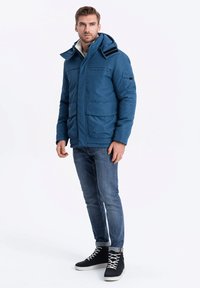 Modrá zateplená bunda s kapucí, která má dvě přední kapsy a proužkované akcenty na ramenou, nošená s modrými džíny a černými vysokými teniskami.