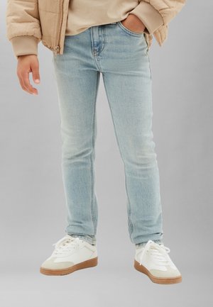 Person trägt hellblaue Skinny-Jeans, beige Daunenjacke, cremefarbenes Hemd und weiße Sneaker mit braunen Sohlen, steht vor grauem Hintergrund.