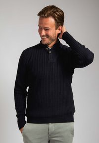 Glimlachende man met lichtbruin haar, gekleed in een zwarte kabelgebreide trui en lichtgrijze broek, met de hand achter het hoofd, tegen een effen achtergrond.