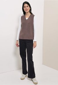 Braunes Strickweste mit V-Ausschnitt, darüber ein langärmeliges weißes Hemd getragen. Kombiniert mit schwarzen Schlaghosen und weißen Turnschuhen.