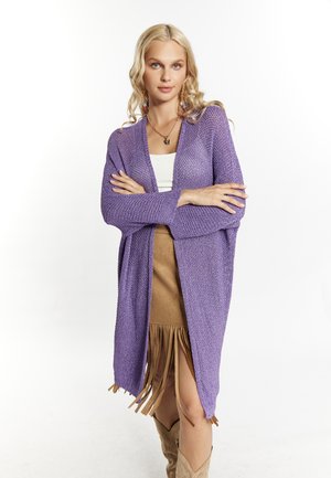 Lila opengehaakte lange cardigan met lange mouwen, gedragen over een witte top en een bruine franjesrok, gecombineerd met tan kniehoge laarzen.