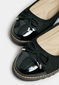 Svarta lägenheter med en glansig lacktå, textilövre, rosettdetalj och en kontrasterande vit söm längs sulan.