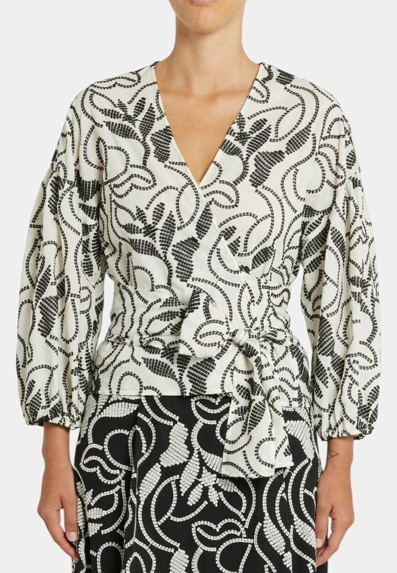 Donna che indossa una blusa avvolgente con motivo astratto bianco e nero e maniche a sbuffo, annodata in vita, abbinata a una gonna con motivo coordinato.