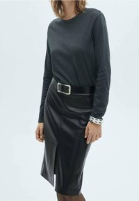 Femme portant un haut à manches longues sombre, une jupe en cuir noire longueur genou avec une fente à l'avant, une ceinture noire avec une boucle argentée et des bracelets argentés au poignet.