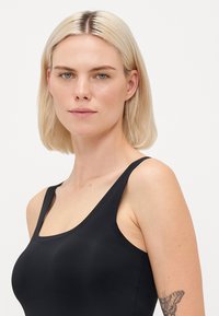 Blonde woman with blue eyes wearing a black sleeveless top, showing a butterfly tattoo on her upper left arm, neutral background.
