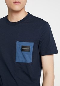 Námořnické bavlněné tričko s kontrastní modrou kapsou na hrudi, na které je černá štítek s logem Calvin Klein. Krátké rukávy a kulatý výstřih.